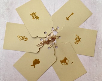 May include: Six beige gift tags with gold foil designs. The designs include a key, a fox, a swirl, a mermaid, a leaf, and a teapot.