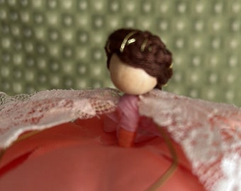 May include: A small doll with brown hair and a pink dress sits on a pink and white fabric background.