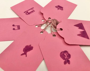May include: Six pink gift tags with different designs. The designs include a crown, a mountain range, a musical note, a sled, a rose, and a silhouette of a woman's head.