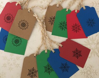 May include: A collection of colorful gift tags in red, blue, green, and brown. Each tag features a black snowflake design and is strung with gold and white twine. The tags are arranged on a white surface with a snowflake pattern.
