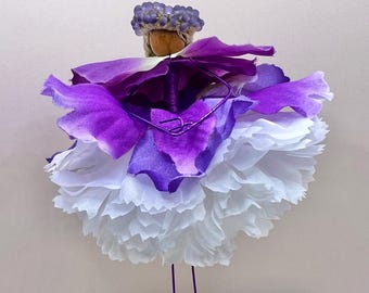 May include: A whimsical floral sculpture featuring a layered design of white and purple petals, resembling a dress. The sculpture has a small, round head with a cluster of purple beads and thin wire legs. The overall aesthetic is delicate and artistic.