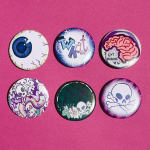 Può includere: Sei spille rotonde con varie illustrazioni: un bulbo oculare blu, il testo "what" in blu e rosa, un cervello rosa con il testo "I EAT BRAINS", un teschio con un arcobaleno, un teschio con foglie verdi e un teschio con ossa incrociate.