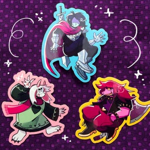 May include: Three die-cut vinyl stickers featuring cartoon characters from the game Deltarune. One sticker shows a dark-haired figure with a sword and cape, another depicts a white-furred creature in a green robe, and the third shows a pink-skinned character with a large axe.