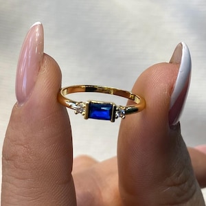 May include: A gold ring featuring a rectangular, deep blue gemstone at the center, flanked by two smaller, clear stones. The ring's band is thin and polished, reflecting light. A delicate piece of jewelry.