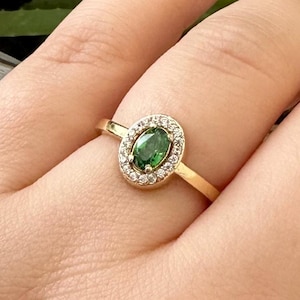 Può includere: Un anello d'oro con una pietra verde ovale, circondata da piccole pietre trasparenti. L'anello è montato su una sottile fascia d'oro. L'anello è mostrato sulla mano di una persona.