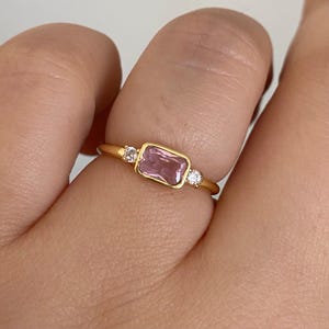 May include: A gold ring featuring a rectangular, light pink gemstone, flanked by two smaller, round, clear gemstones. The ring is set in a gold band, showcasing a minimalist design. The ring is shown on a finger.