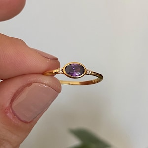 Può includere: Un anello d'oro con una pietra preziosa ovale, viola intenso. L'anello ha una fascia sottile ed è impreziosito da due piccole pietre trasparenti su entrambi i lati della pietra principale. L'anello è tenuto dalle dita di una persona.