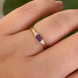 Op de afbeelding: Een gouden ring met een rechthoekige, dieppaarse edelsteen, geflankeerd door twee kleinere, ronde, heldere stenen. De dunne gouden band van de ring creëert een delicaat ontwerp. De ring wordt op een vinger getoond.