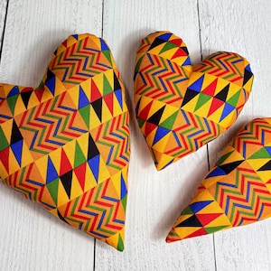 May include: Three heart-shaped decorative pillows with a vibrant geometric pattern in yellow, red, green, blue, and black. The pillows are arranged on a white wooden surface, showcasing their colorful design.