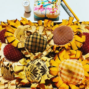 May include: A close-up of a handmade sunflower wreath with multiple layers of yellow fabric petals and various patterned fabric centers. The wreath is made with a variety of fabrics, including plaid, gingham, and floral prints.