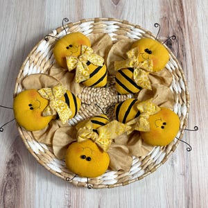 May include: Six yellow felt bumblebees with black stripes and yellow and white floral bows are arranged in a woven basket.