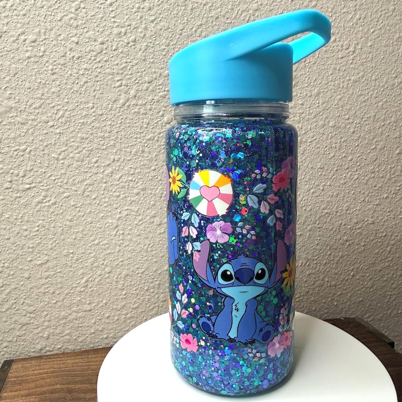 Peut inclure: Une bouteille d'eau transparente avec un couvercle bleu et un motif de personnage de dessin anim&eacute;. La bouteille est remplie de paillettes bleues et d&eacute;cor&eacute;e de fleurs et d'un personnage de dessin anim&eacute;. Le couvercle a une poign&eacute;e.
