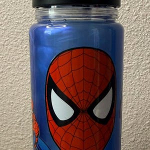 May include: A blue translucent water bottle featuring a large graphic of the Spider-Man mask with a red and black web pattern and white eyes. The bottle has a black screw-top lid with an integrated carrying handle and a clear plastic rim.
