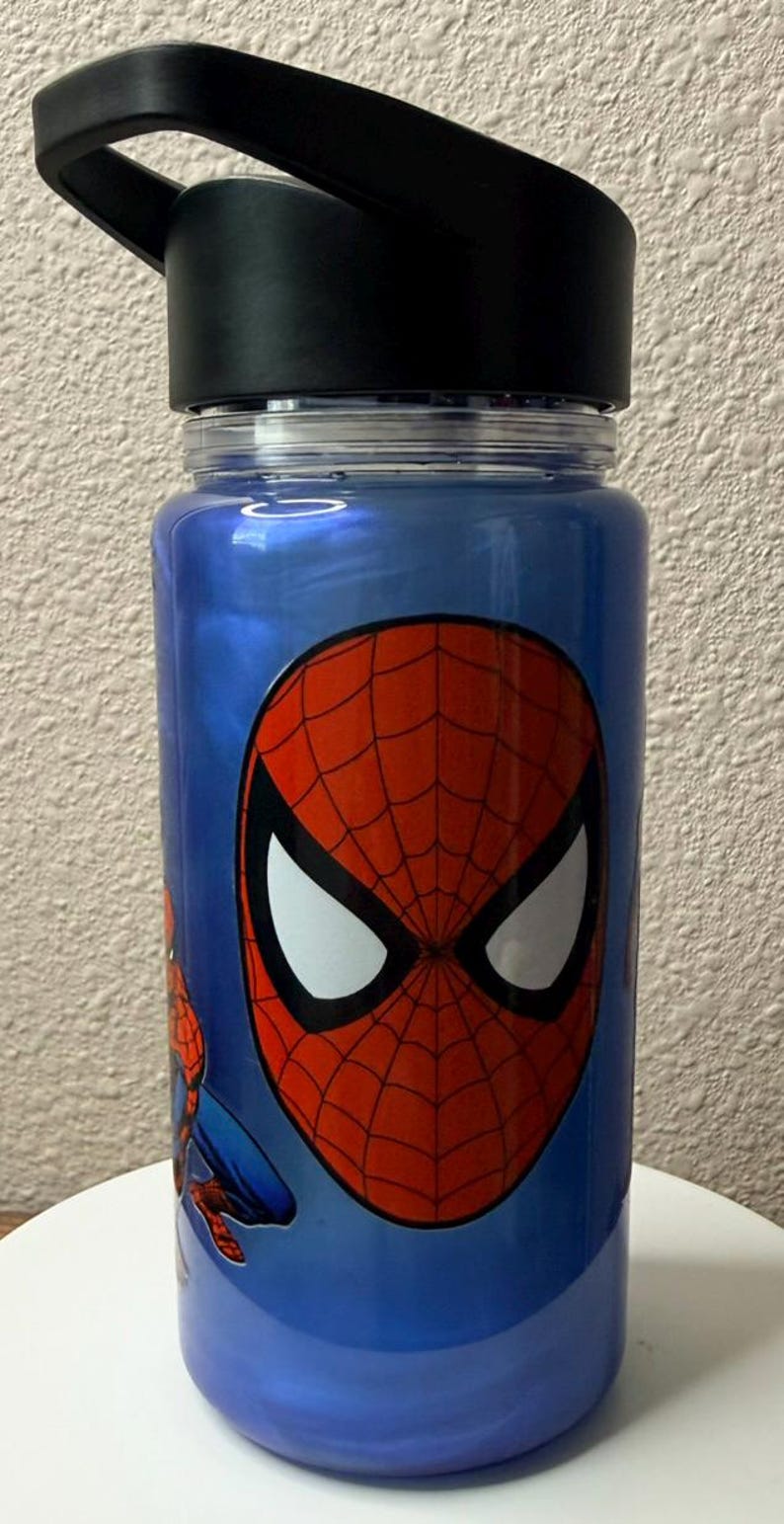 May include: A blue translucent water bottle featuring a large graphic of the Spider-Man mask with a red and black web pattern and white eyes. The bottle has a black screw-top lid with an integrated carrying handle and a clear plastic rim.