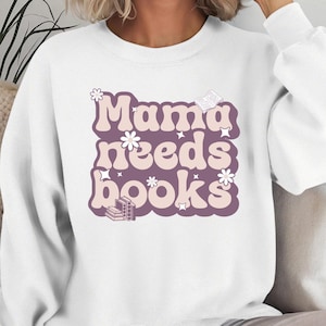 May include: A white long-sleeve crewneck sweatshirt featuring a graphic print with the text Mama needs books in a retro font. The design includes light pink and purple lettering, small white flower icons, star shapes, and a stack of books.