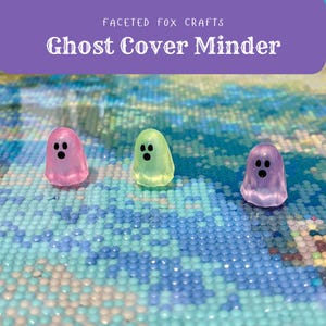 May include: Three translucent ghost-shaped cover minders in pink, green, and purple, each with three black dots for eyes and mouth. The text "Ghost Cover Minder" is displayed above the ghosts. The background is a mosaic of blue and white beads.