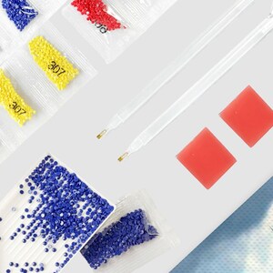 May include: An assortment of diamond painting supplies, including packets of blue, red, and yellow diamond beads, labeled with the number "307". Also included are two clear applicator pens and two square red wax pads, all arranged on a white surface.