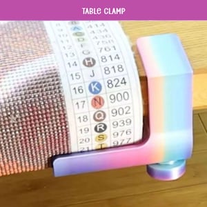 May include: A multi-colored table clamp, with a pastel rainbow gradient, holds a diamond painting project in place. The clamp is made of plastic and has a sturdy design. The text "TABLE CLAMP" is visible at the top.