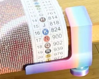 May include: A gradient-colored table clamp with a smooth finish, securing a diamond painting canvas. The canvas features a printed legend with numbers and letters, including symbols for colors 814, 818, 824, 900, 902, 939, 976, and 977. The clamp has a rounded base.