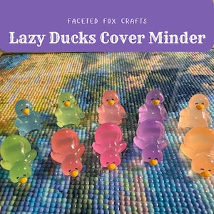 May include: A selection of translucent duck-shaped cover minders in shades of blue, green, pink, and purple, each with a yellow beak. The text "Lazy Ducks Cover Minder" is displayed above the ducks. These are used for diamond painting.