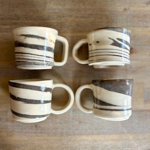 May include: Four ceramic mugs with a cream-colored base and dark brown, horizontal stripe designs. Each mug has a rounded handle and a slightly textured surface. The mugs are arranged in a square formation on a wooden surface.