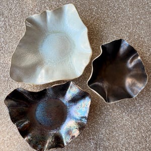 May include: Three ceramic bowls with irregular, ruffled edges. One bowl is a glossy off-white with a textured surface. The second bowl is a dark, metallic brown. The third bowl features a deep, iridescent finish with blue and bronze tones.