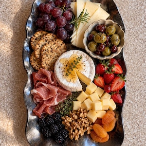 May include: A dark metallic wavy-edged serving platter holds a charcuterie arrangement featuring red grapes, sliced cheese, crackers, olives in a small bowl, brie with honey, prosciutto, strawberries, cubed cheese, walnuts, and dried apricots.