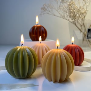 May include: Four spherical, ribbed candles in olive green, light beige, golden yellow, and deep red, each with a lit wick. The candles are arranged in a cluster, displaying a vertical fluted texture that creates a rounded, pumpkin-like shape.
