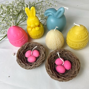 Peut inclure: Une collection de bougies sur le th&egrave;me de P&acirc;ques de diff&eacute;rentes formes et couleurs. Comprend une bougie lapin jaune, une bougie en forme de lapin bleu, des bougies en forme d'&oelig;uf roses et jaunes, et deux bougies nid marron avec des &oelig;ufs roses.