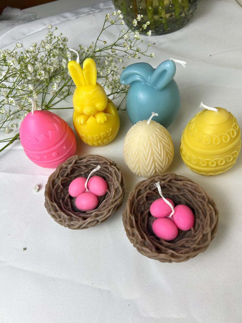 Peut inclure: Une collection de bougies sur le th&egrave;me de P&acirc;ques de diff&eacute;rentes formes et couleurs. Comprend une bougie lapin jaune, une bougie en forme de lapin bleu, des bougies en forme d'&oelig;uf roses et jaunes, et deux bougies nid marron avec des &oelig;ufs roses.