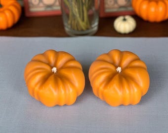 May include: Two orange pumpkin-shaped candles with white wicks sit on a light blue surface. The candles are in the foreground, with other pumpkins and a vase of flowers in the background. The scene evokes a fall or autumn theme.