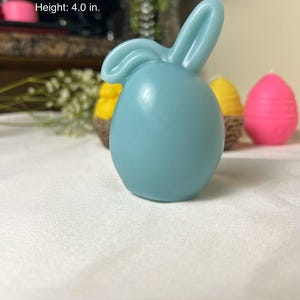 Peut inclure: Une bougie en forme de lapin bleu clair, d'environ 6,35 cm de large et 10,16 cm de haut, avec une m&egrave;che blanche. D'autres bougies en arri&egrave;re-plan comprennent des bougies en forme d'&oelig;uf jaunes et roses. Les bougies sont sur une surface blanche.
