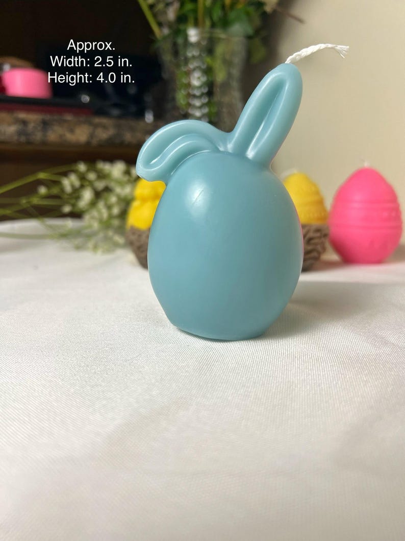 Peut inclure: Une bougie en forme de lapin bleu clair, d'environ 6,35 cm de large et 10,16 cm de haut, avec une m&egrave;che blanche. D'autres bougies en arri&egrave;re-plan comprennent des bougies en forme d'&oelig;uf jaunes et roses. Les bougies sont sur une surface blanche.