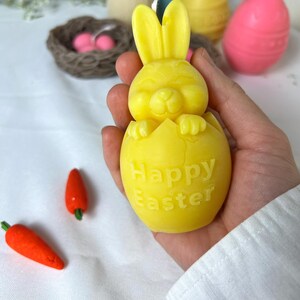 Peut inclure: Une bougie jaune en forme de lapin sortant d'un &oelig;uf, avec les mots "Happy Easter" sur l'&oelig;uf. Deux bougies en forme de carottes oranges et d'autres bougies sur le th&egrave;me de P&acirc;ques sont en arri&egrave;re-plan.