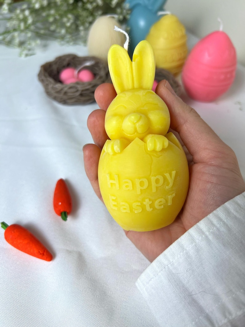 Peut inclure: Une bougie jaune en forme de lapin sortant d'un &oelig;uf, avec les mots "Happy Easter" sur l'&oelig;uf. Deux bougies en forme de carottes oranges et d'autres bougies sur le th&egrave;me de P&acirc;ques sont en arri&egrave;re-plan.