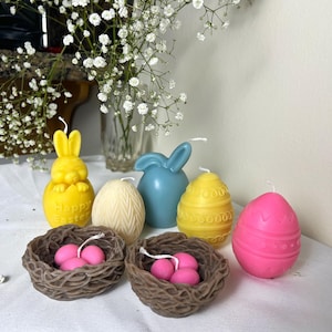 Peut inclure: Diverses bougies sur le th&egrave;me de P&acirc;ques, dont un lapin jaune, un lapin bleu et des bougies en forme d'&oelig;uf jaunes et roses. Deux nids bruns contiennent des bougies en forme d'&oelig;ufs roses. Des fleurs blanches sont en arri&egrave;re-plan.