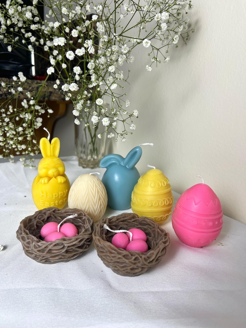 Peut inclure: Diverses bougies sur le th&egrave;me de P&acirc;ques, dont un lapin jaune, un lapin bleu et des bougies en forme d'&oelig;uf jaunes et roses. Deux nids bruns contiennent des bougies en forme d'&oelig;ufs roses. Des fleurs blanches sont en arri&egrave;re-plan.