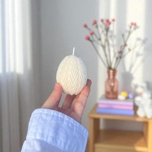 Peut inclure: Une bougie cr&egrave;me en forme d'&oelig;uf avec un motif en chevrons textur&eacute;. La bougie est tenue dans une main, avec une manche de chemise blanche visible. Une petite m&egrave;che blanche d&eacute;passe du haut. L'arri&egrave;re-plan comprend un vase de fleurs roses.