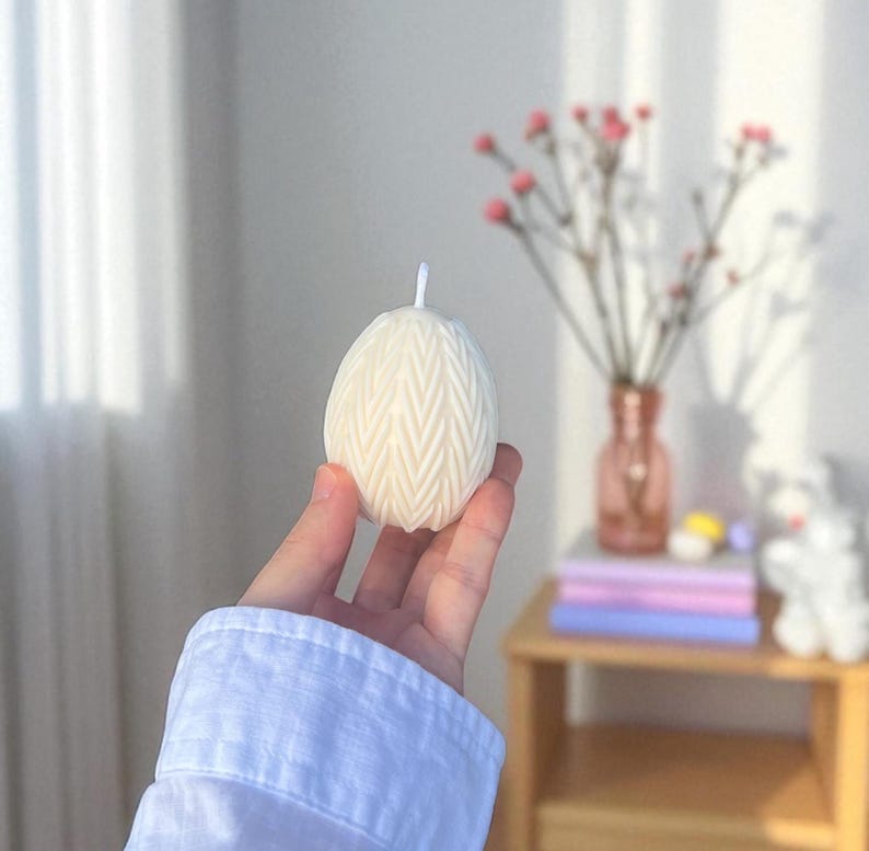 Peut inclure: Une bougie cr&egrave;me en forme d'&oelig;uf avec un motif en chevrons textur&eacute;. La bougie est tenue dans une main, avec une manche de chemise blanche visible. Une petite m&egrave;che blanche d&eacute;passe du haut. L'arri&egrave;re-plan comprend un vase de fleurs roses.
