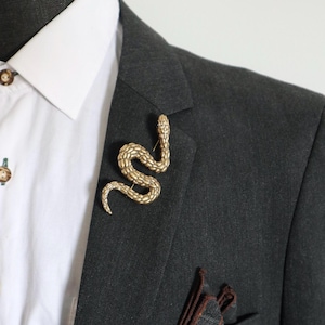Op de afbeelding: Een goudkleurige slangenbroche met een gestructureerd, schubachtig oppervlak, opgespeld op de revers van een donker antracietgrijze blazer. De broche heeft een kronkelende S-vorm en is voorzien van een gedetailleerde kop en een dunne staart.