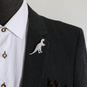 Pode incluir: Um broche de dinossauro prateado coberto com pequenos cristais transparentes, preso na lapela de um blazer cinza-carvão escuro sobre uma camisa branca com botões castanhos. A figura do dinossauro é representada em perfil.
