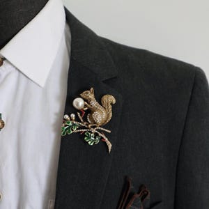 Op de afbeelding: Een goudkleurige eekhoornbroche met heldere strass-steentjes, een witte kunstparel en groene geëmailleerde eikenbladeren op een tak. De eekhoorn houdt een witte kunstparel vast, met extra kleine parels die de takstructuur accentueren.