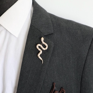 Könnte beinhalten: Eine goldfarbene Schlangenbrosche mit strukturierter Oberfläche und eingelassenen klaren Steinen, befestigt am Revers eines dunkel anthrazitfarbenen Blazers über einem weißen Hemd mit Kragen. Die Schlange ist in einer geschwungenen S-Form dargestellt.