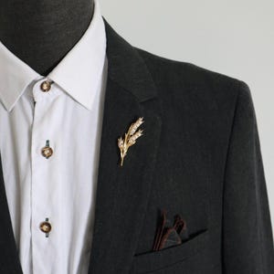 Könnte beinhalten: Eine goldfarbene Weizenähren-Brosche am Revers eines dunkel anthrazitfarbenen Blazers. Der Blazer wird über einem weißen Hemd mit braunen Knöpfen getragen. Ein dunkelbraunes Einstecktuch steckt in der Brusttasche des Blazers.