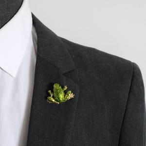 May include: A bright green frog-shaped brooch with a glossy finish and metallic gold-toned edges. The frog is depicted in a seated position with its legs tucked underneath its body, featuring textured skin details across its back and head.