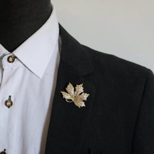 Könnte beinhalten: Eine goldfarbene Ahornblatt-Brosche mit strukturierter Oberfläche und eingebetteten klaren Kristallen, befestigt am Revers eines dunkel anthrazitfarbenen Blazers über einem weißen Hemd mit dunkelbraunen Knöpfen.