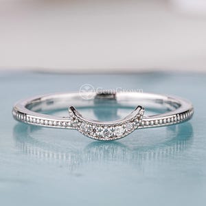 May include: A silver-colored ring with a crescent moon design. The ring features small, clear gemstones set in the crescent and along the band. The ring is displayed on a light blue surface.