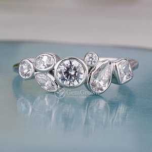 May include: A silver ring featuring a cluster of various sized and shaped clear gemstones. The ring is displayed on a light blue surface. The words 'GemGrow USA' are printed on the surface.