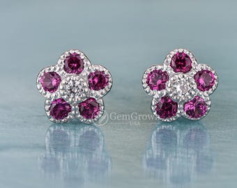 May include: A pair of flower-shaped stud earrings. Each earring features five deep pink gemstones surrounding a central cluster of smaller, clear stones. The earrings are set in a silver-colored metal and rest on a light blue surface.