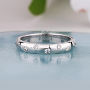 May include: A polished silver ring with a series of small, round, clear gemstones set into the band. The ring is displayed on a light blue surface, with a blurred white flower in the background. The ring has a smooth, reflective surface.